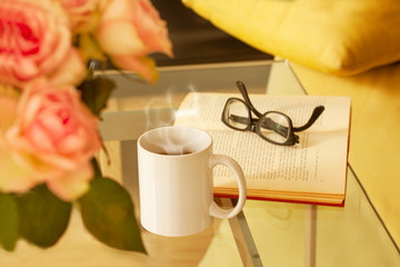 Book, glasses, roses and a mug with tea on modern glass table. Living room interior home decor concept.