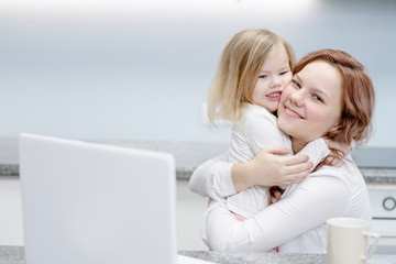 Happy young mother hugs her little daughter at home in the kitchen near an open laptop