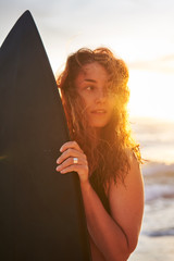 Slim woman in bikini carrying surfboard and walking on wet sand towards waving sea on beach