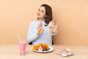 Teenager girl eating waffles isolated on beige background nervous stretching hands to the front