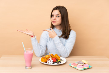 Teenager girl eating waffles isolated on beige background holding copyspace imaginary on the palm to insert an ad