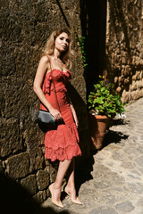 Vogue style elegant portrait of beautiful fashion woman wavy shine blonde long hair. Model in pink orange coral dress with bright makeup in old town medieval Italy