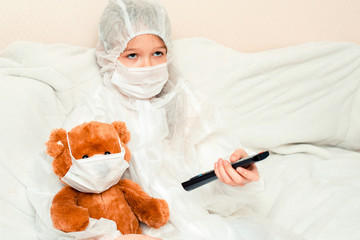 Young girl with teddy bear, in masks and a white medical gown, lie on bed and watch TV, Concept quarantine, stay at home