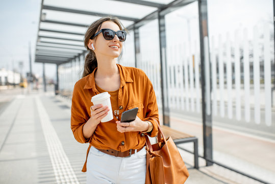 Lifestyle portrait of a young and cheerful woman standing with phone and coffee cup on the public transport stop outdoors. Urban business travel and transportation concept
