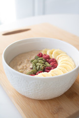 Homemade healthy breakfast: oatmeal porridge with banana, ripe raspberries and pumpkin seeds with ingredients in the background