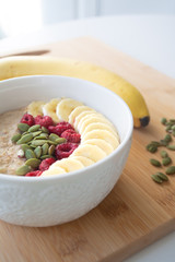Homemade healthy breakfast: oatmeal porridge with banana, ripe raspberries and pumpkin seeds with ingredients in the background