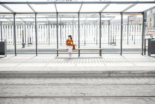 Woman sitting alone at the public transport stop on a sunny day outdoors, general plan on a modern stop. Concept of a transportation and urban life