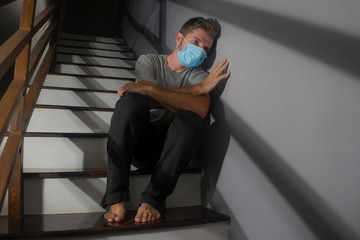 dark portrait of young scared and worried man in protective mask sitting on stairs at home staircase during lockdown and quarantine for covid-19 virus pandemic in dramatic light