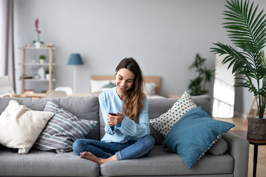 Online chat. Smiling woman talking on smartphone with friends while sitting on sofa at home.