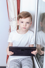 Distance learning in self-isolation mode and quarantine concept. A seven-year-old boy sits by the window and holds a tablet in his hands. Online school, video conferencing lessons.