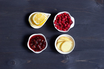 Raspberry jam, fresh cranberries, slices of ginger and lemon in white bowls as healthy organic treatment on black wooden background 