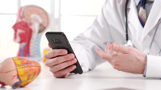 A medical professional doctor wearing a white lab coat and a stethoscope is having a video chat call with his patient, using a smartphone mobile devise app from his office