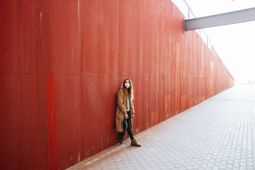 a young girl in a brown coat with a medical mask on her face,fighting against the corona 19 virus infection on the street, near the red wall