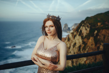 Beautiful red-haired girl in a gold dress and crown stands on the shore of the ocean