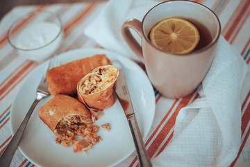 Stuffed pancakes on a round white plate, next to a glass of tea with lemon and sour cream in a small round plate