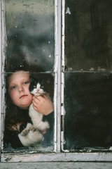Portrait of little plump girl with her lovely kitten looking out of old window in village house.