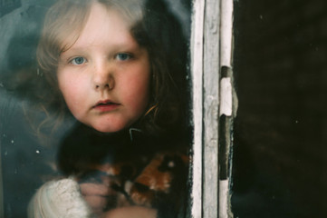 Portrait of a little girl with tranquil face expression looking out of old dirty window with reflections.