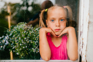 Little cute girl with blond hair and pink dress with beautiful flowers behind the window