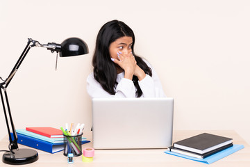 Student asian girl in a workplace with a laptop isolated on beige background covering mouth and looking to the side