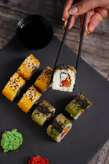 Sushi set on a square black plate of stone. A  hand holds a roll of chopsticks. View from above.  Wood Background