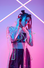 Beautiful asian woman in a raincoat listens music in headphones around colourful neon