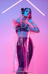 Beautiful asian woman in a raincoat listens music in headphones around colourful neon