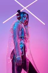 Tuinposter Beautiful asian woman in a raincoat listens music in headphones around colourful neon © micro