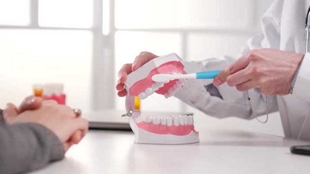 Doctor of dental surgery in a lab coat and stethoscope explaining to the patient the right way of brushing teeth by demonstrating it on an anatomical model of a human mouth