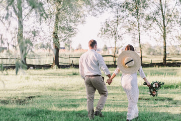 Wedding. The groom in a white shirt and light pants holds the bride s hand in a beautiful dress and a straw hat with a beautiful wedding bouquet in his hands. back in the frame