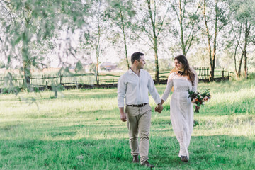 Wedding. The groom in a white shirt and light pants holds the bride s hand in a beautiful dress and a straw hat with a beautiful wedding bouquet in his hands. go to meet the camera