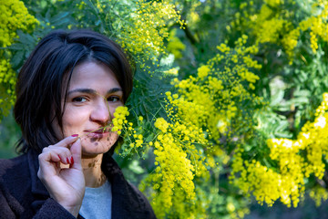 Pretty smiling brunette near a large bush of Yellow fluffy mimosa with green leaves