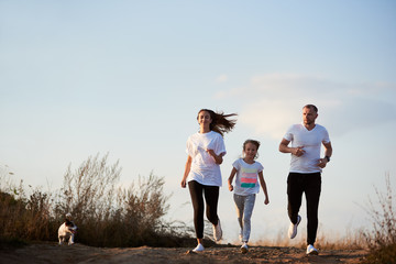 Young and beautiful family - father, mother and daughter are jogging with their dog outside the city on the village road on the setting sun