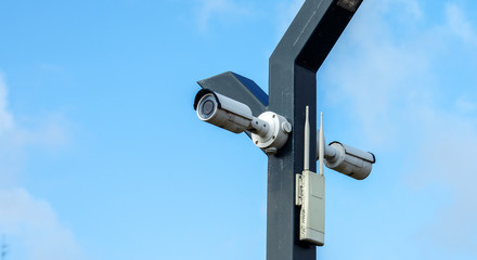 Two cctv cameras and a wifi antenna hang on a pole against the sky