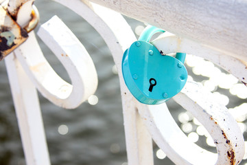 A blue or turquoise heart-shaped castle is fastened to a white fence and hangs against a background of shiny water