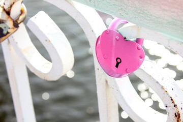 A pink heart-shaped padlock hanging on a white fence against a water background with bokeh and...