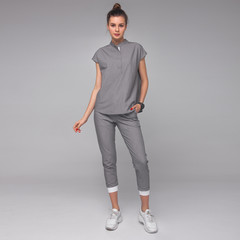 young stylish brunette nurse with long legs in gray medical costume is standing with hand in her pocket, looking straight and smiling cute on gray wall background. medical concept. free space