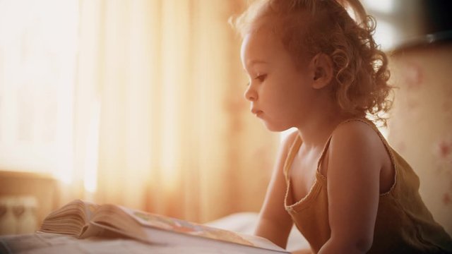 Peaceful baby girl looks at pictures in the book in sunny bedroom at home