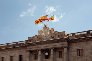 View of Spanish and Catalan flags waving at city square called "Placa de Sant Jaume" in "Ciutat Vella" district (Gothic Quarter) in Barcelona. It is a sunny summer day.