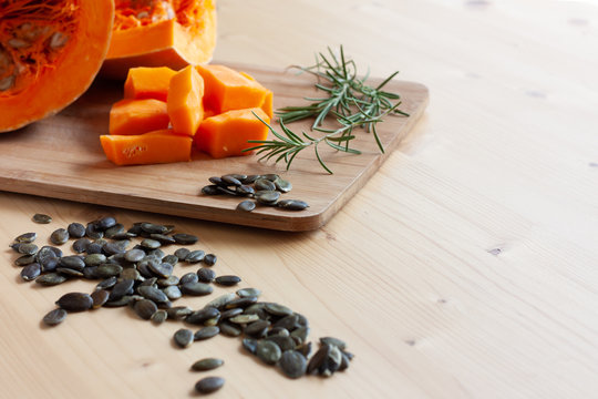 Red pumpkin cut into cubes on wooden cutting borad with pampkin seeds and rosemary on wooden light background. Space for text. Horizontal view.
