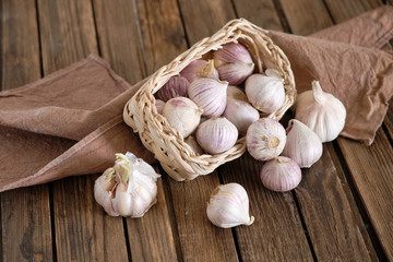 White round garlic falls from a wicker basket on a brown wooden background with a dark napkin. Close-up photo