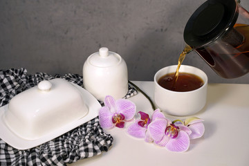 Fototapeta na wymiar The tea is being poured from the bright glass teapot. Tea set and pink orchids on table