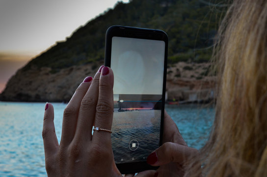 White woman with her mobile phone taking a panoramic picture of the sunset in Benirras Beach in Ibiza
