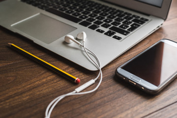 Espacio de tele trabajo. Ordenador portátil con auriculares blancos, lápiz amarillo y negro, teléfono móvil, sobre mesa de madera.