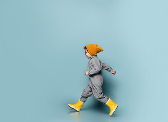 Brunet child in sunglasses, orange hat, gray overall and yellow rubber boots. He is running, posing...