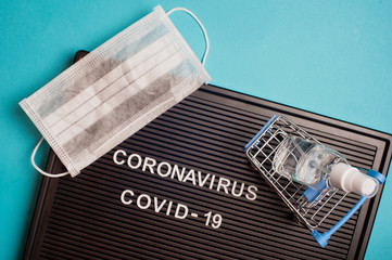 Coronavirus - COVID -19 - text on black letter board, surgical mask and sanitizer gel in little shopping cart on blue background.