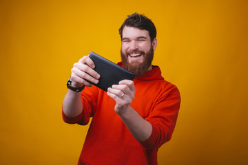 Photo of a cheerful young man is excited about playing games on his tablet on yellow background.