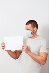 a serious Caucasian man in light clothing and a medical mask on his face holds a blank sheet of paper on a white background. . stay at home