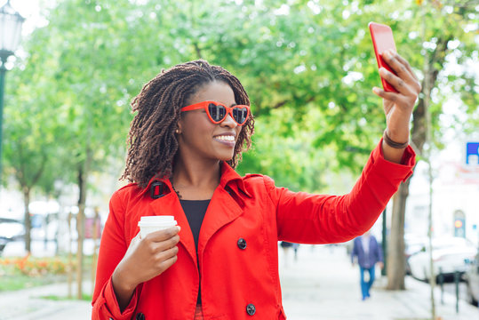 Content woman in sunglasses taking selfie in park. Beautiful smiling young African American woman holding coffee to go and taking selfie with smartphone outdoors. Technology concept