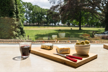 A wooden board with cheese olives tomatoes and wine in front of a window with view on the garden
