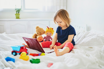 Toddler girl with laptop and toys in bed